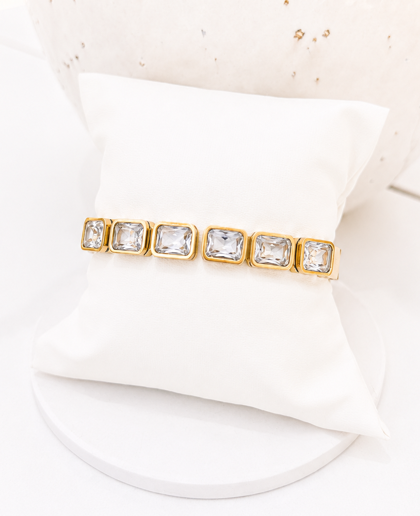 
                  
                    A gold bracelet with five rectangular clear gemstones, displayed on a white fabric jewelry stand.
                  
                
