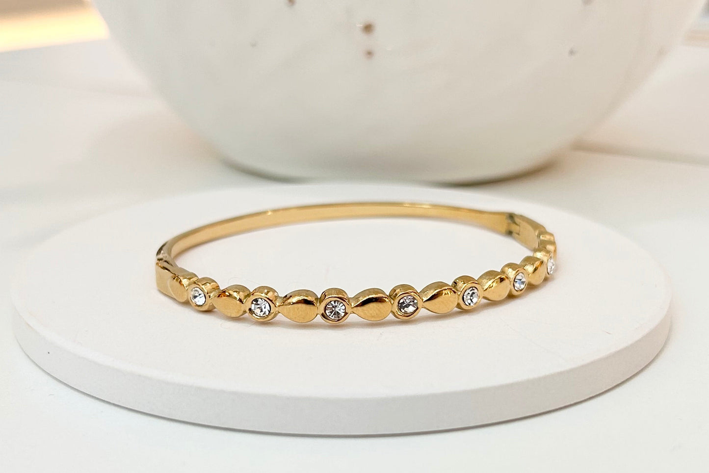 
                  
                    A delicate gold bangle bracelet with a row of small clear stones, displayed on a round white coaster with a white speckled ceramic vase in the background.
                  
                
