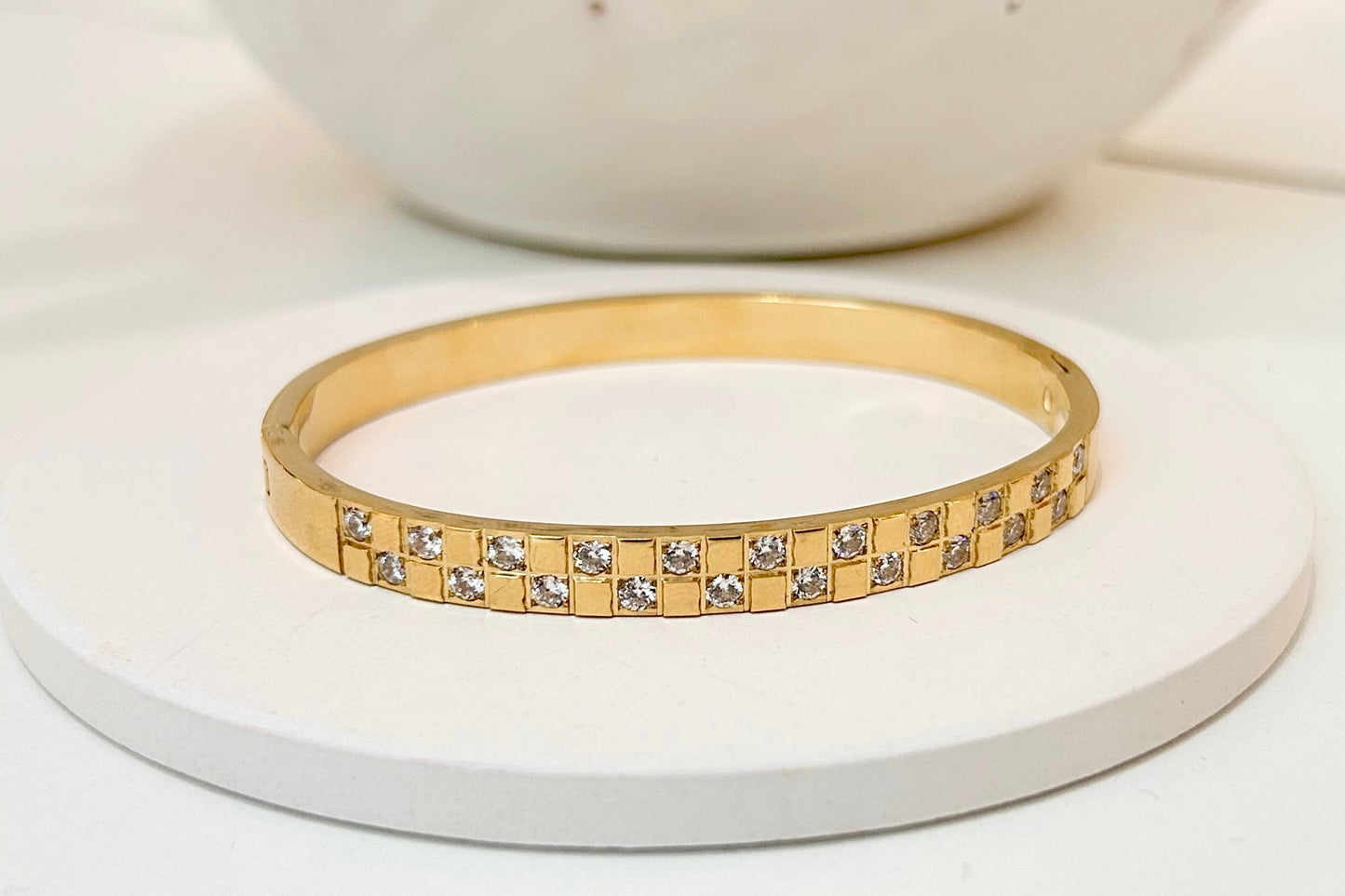 A gold bangle bracelet with a checkered pattern of clear stones, displayed on a round white coaster with a white ceramic bowl in the background.
