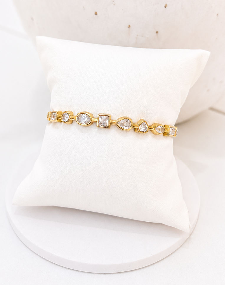 
                  
                    A delicate gold bracelet with clear gemstones displayed on a small white cushion stand, set against a soft-focus white textured background.
                  
                