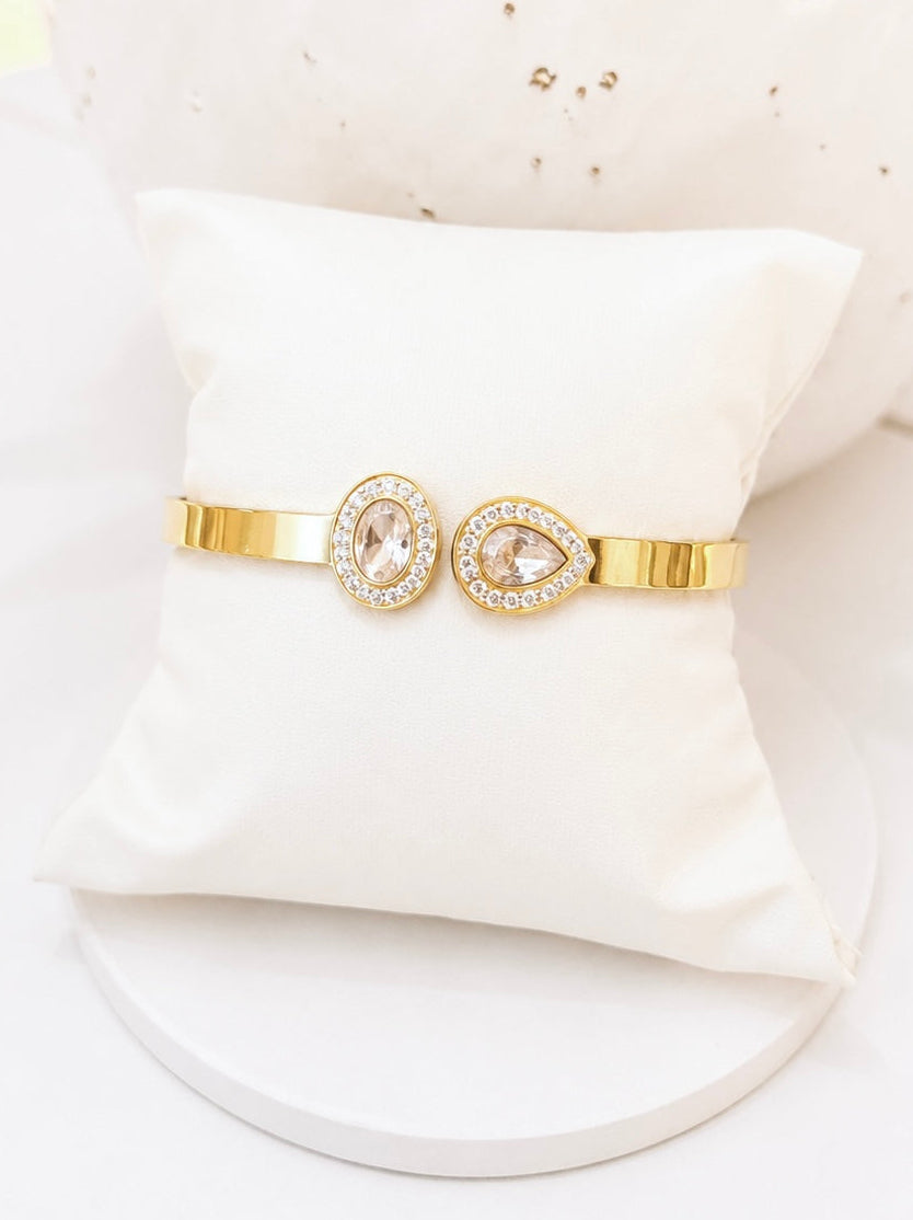 
                  
                    A gold-toned double-band bracelet displayed on a white cushion. The bracelet features two oval bezel-set clear stones surrounded by small clear crystals, with one of the oval stones replaced by a pear-cut clear stone.
                  
                