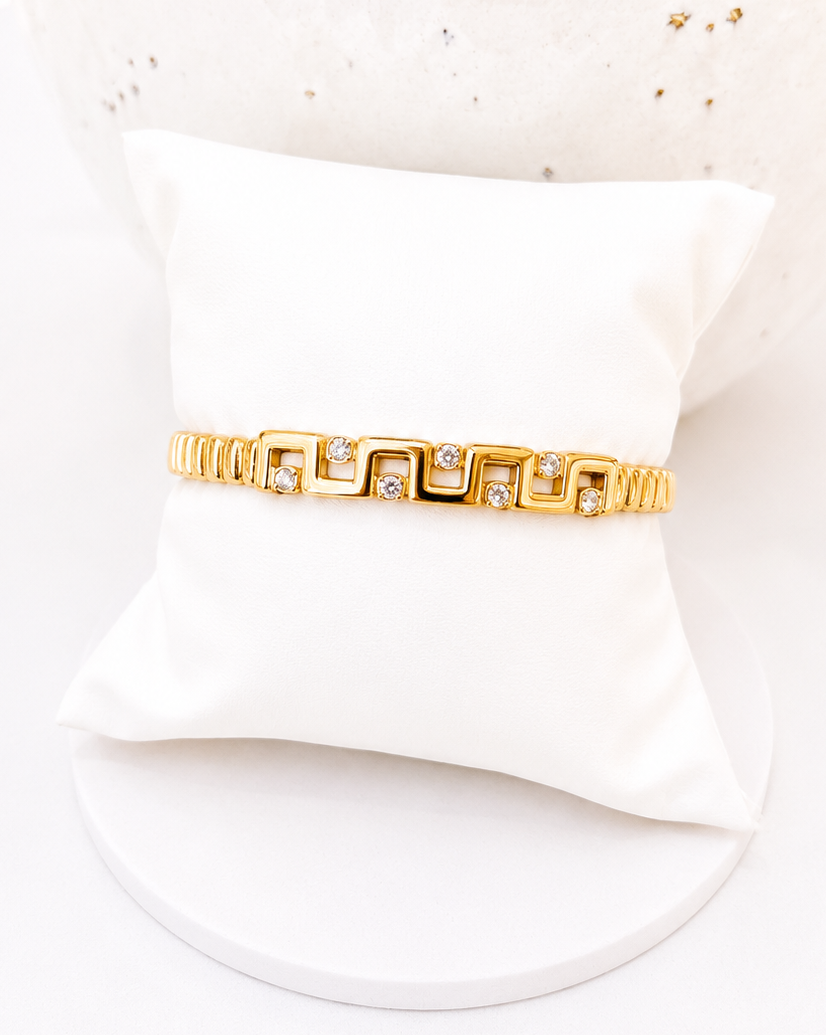 A gold bracelet with a repeating geometric square link design and small clear stones, displayed on a white pillow against a white background.