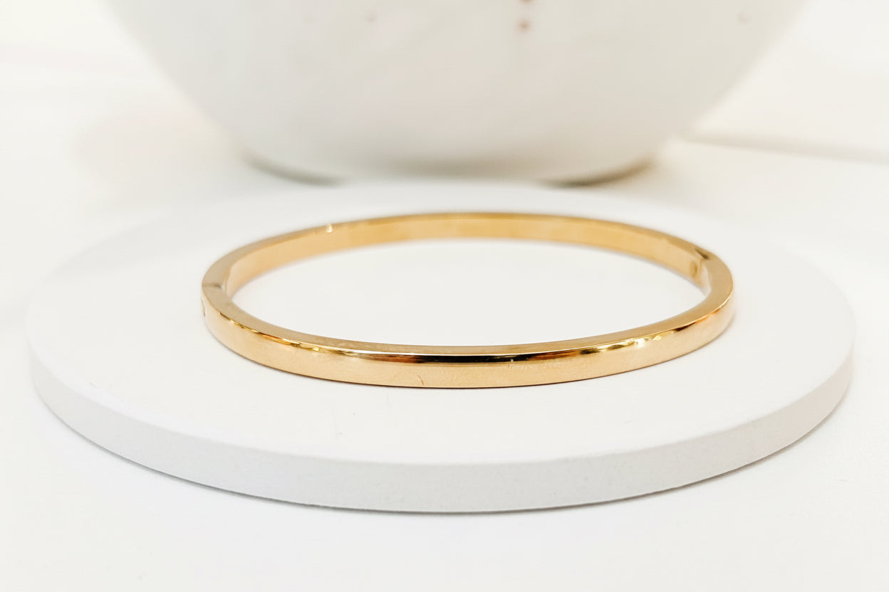 A thin polished gold bangle bracelet displayed on a round white coaster, with a blurred white ceramic bowl in the background.