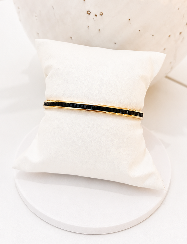 
                  
                    A slim gold bangle bracelet with a black inlay, displayed on a white fabric jewelry pillow against a white background.
                  
                