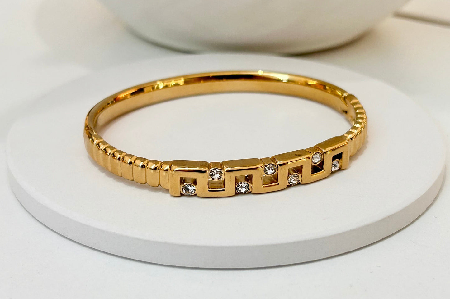 A gold bangle bracelet with geometric links and small clear stones, displayed on a round white pedestal.