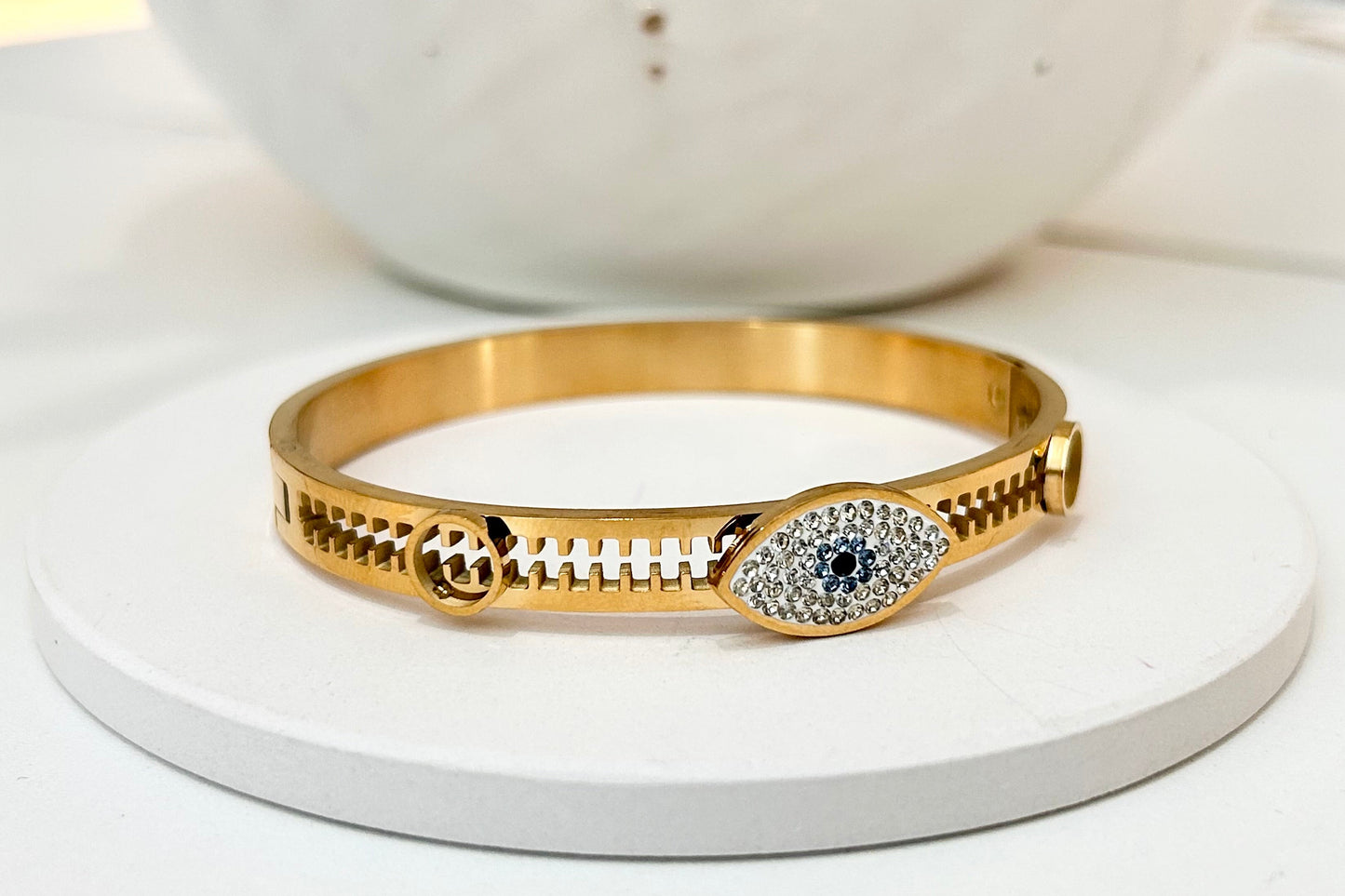 
                  
                    A gold bangle bracelet with a central evil eye motif paved with small clear stones and a dark blue center stone, displayed on a round white stand.
                  
                