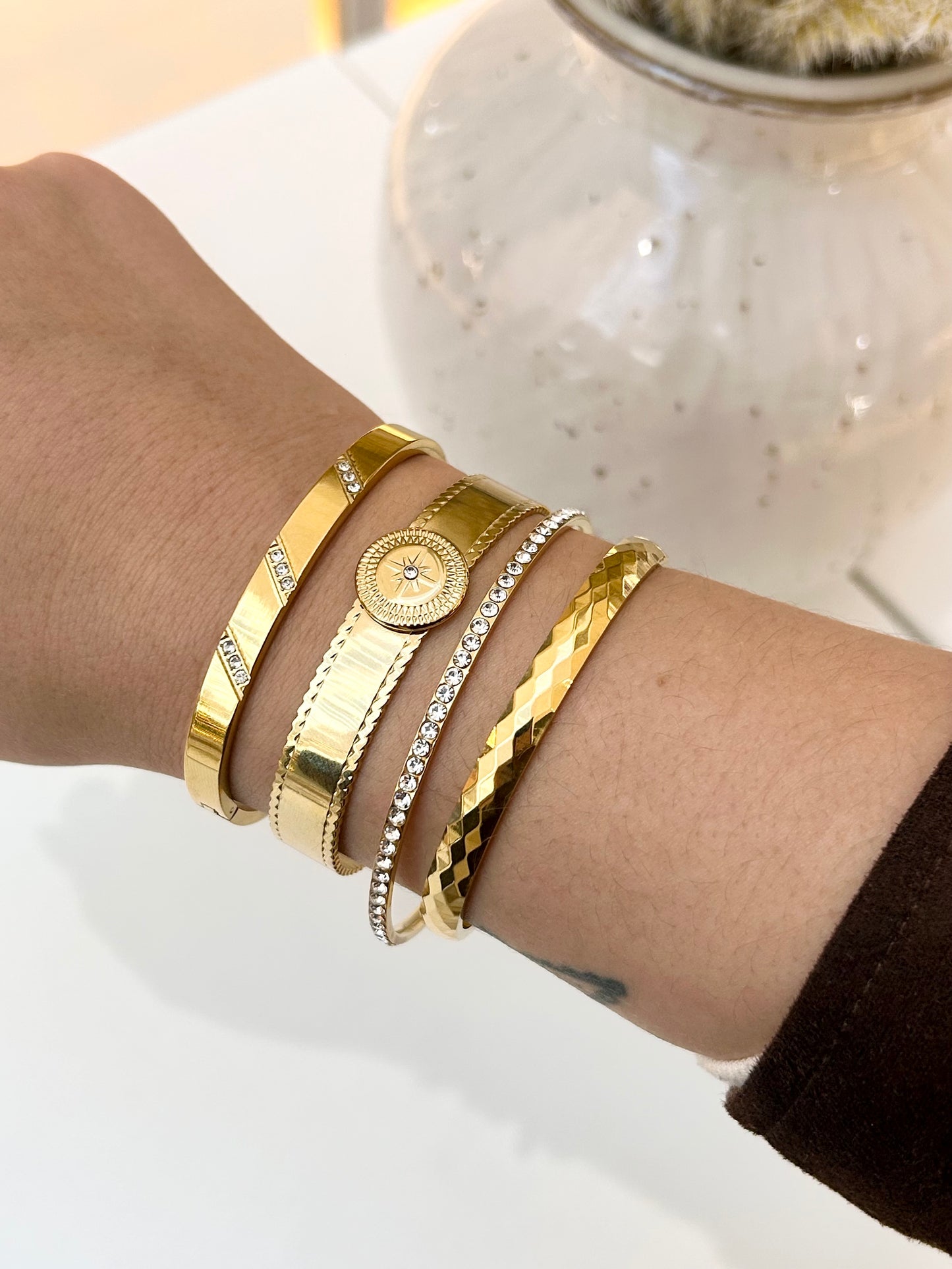 A close-up of a person's wrist wearing five stacked gold-toned bracelets, including a bangle with small clear stones, a textured bangle, a chain-link style bracelet, and a round bezel-set bracelet, against a white background with a decorative vase.