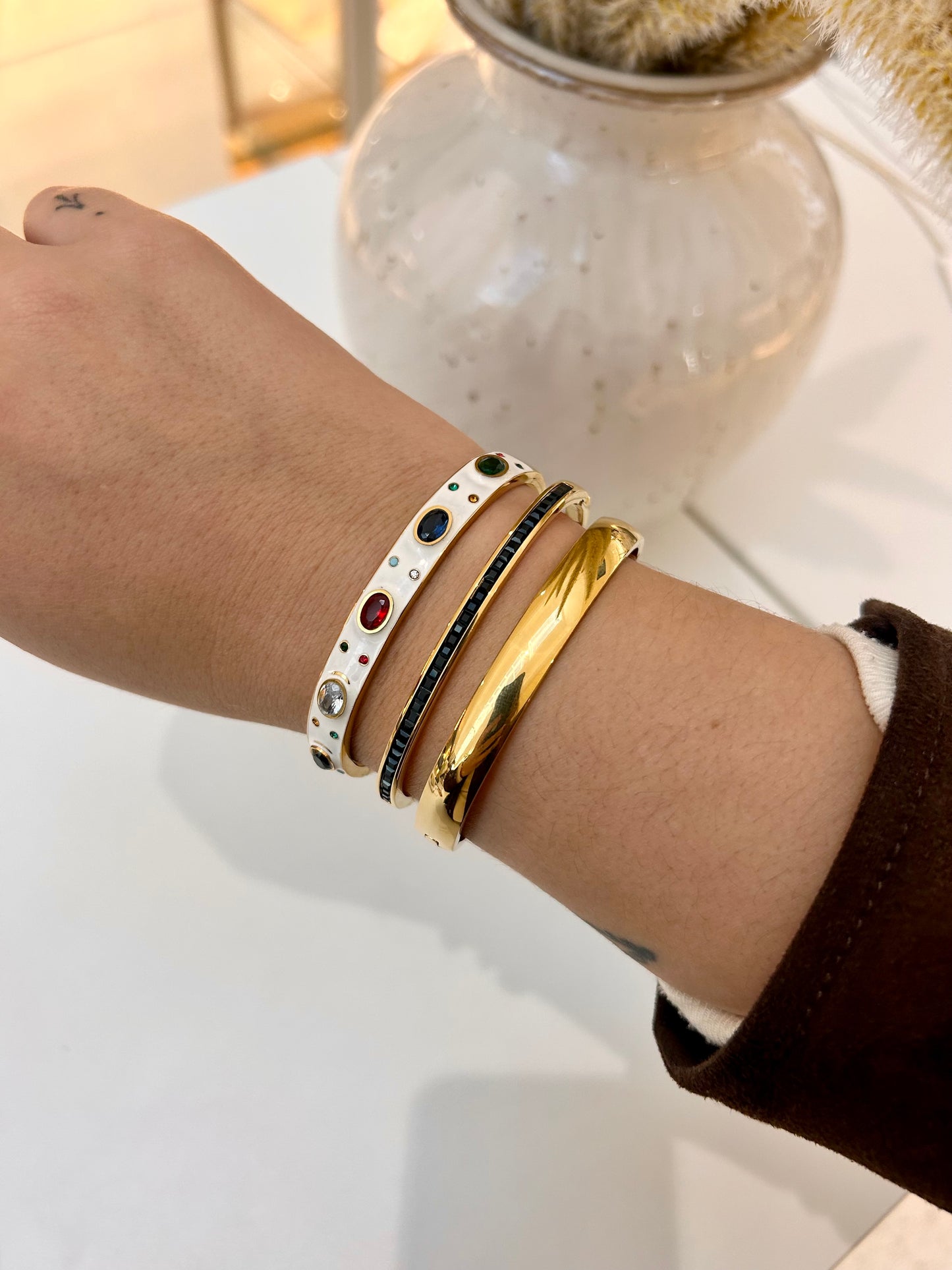 A close-up of a person's wrist wearing three stacked bracelets: a white bangle with colorful rhinestones, a black bangle with red and green stones, and a polished gold bangle.