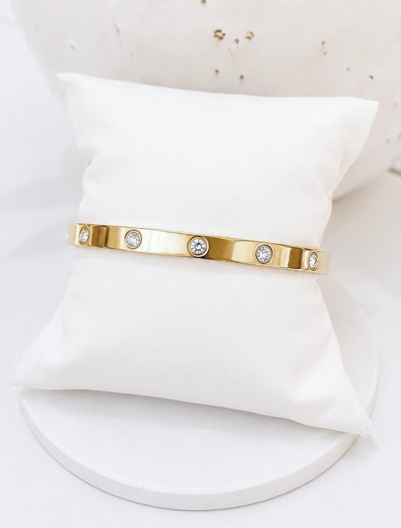 
                  
                    A slim gold bangle bracelet with three clear round stones, displayed on a white fabric pillow against a white background.
                  
                