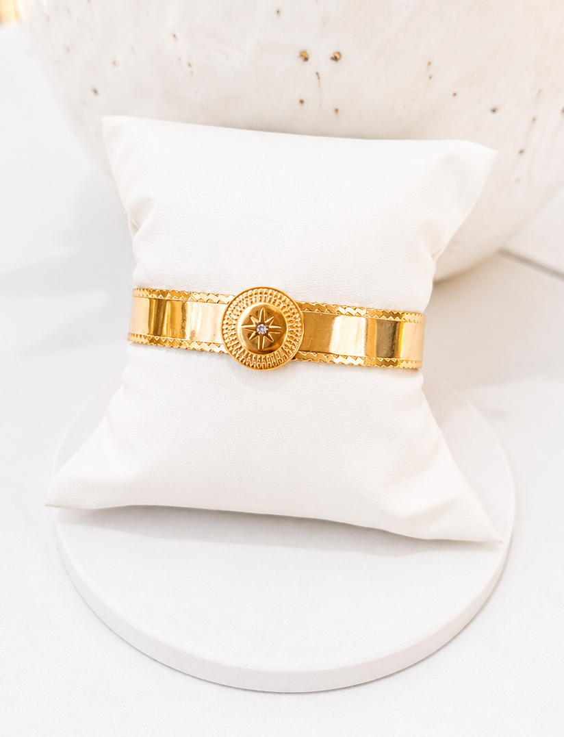 
                  
                    A gold-tone metal bangle bracelet with a circular starburst emblem on the center band, displayed on a small white pillow stand.
                  
                