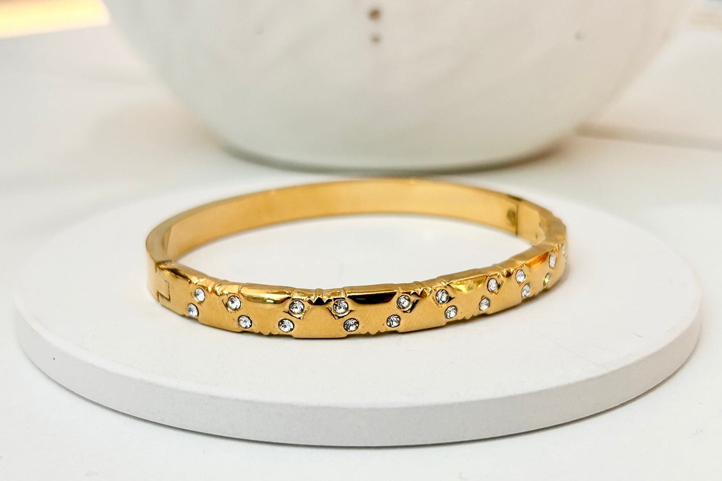 
                  
                    A gold bangle bracelet with small clear rhinestones set along the front, displayed on a round white coaster with a white ceramic bowl in the background.
                  
                