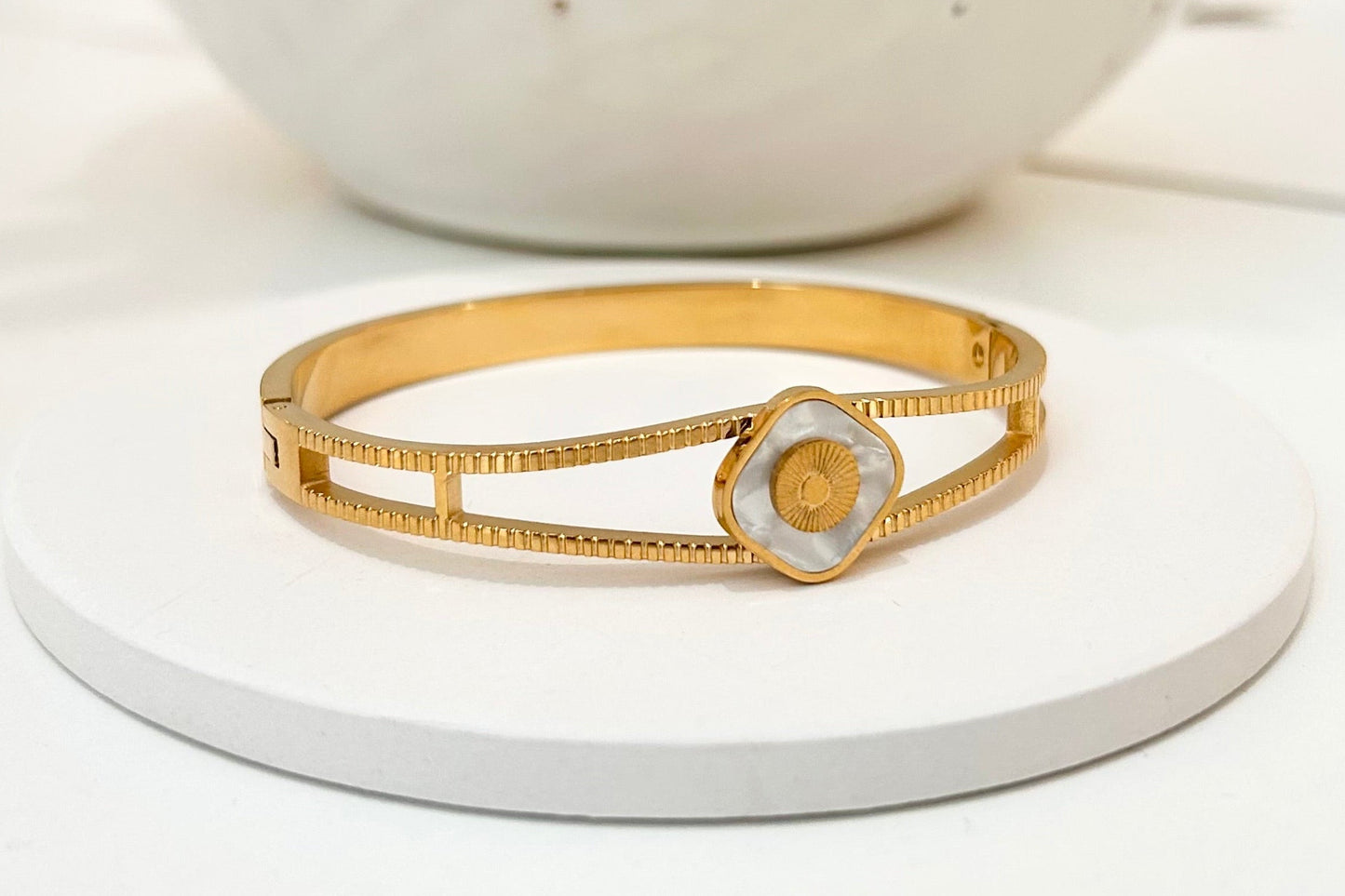 A gold bangle bracelet with a textured floral center, displayed on a white circular stand with a blurred white speckled vase in the background.
