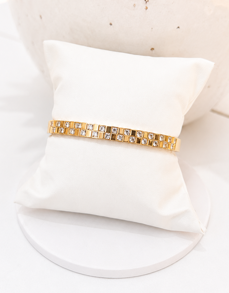 
                  
                    A gold bracelet with square-cut clear stones, displayed on a small white pillow in front of a textured white vase.
                  
                