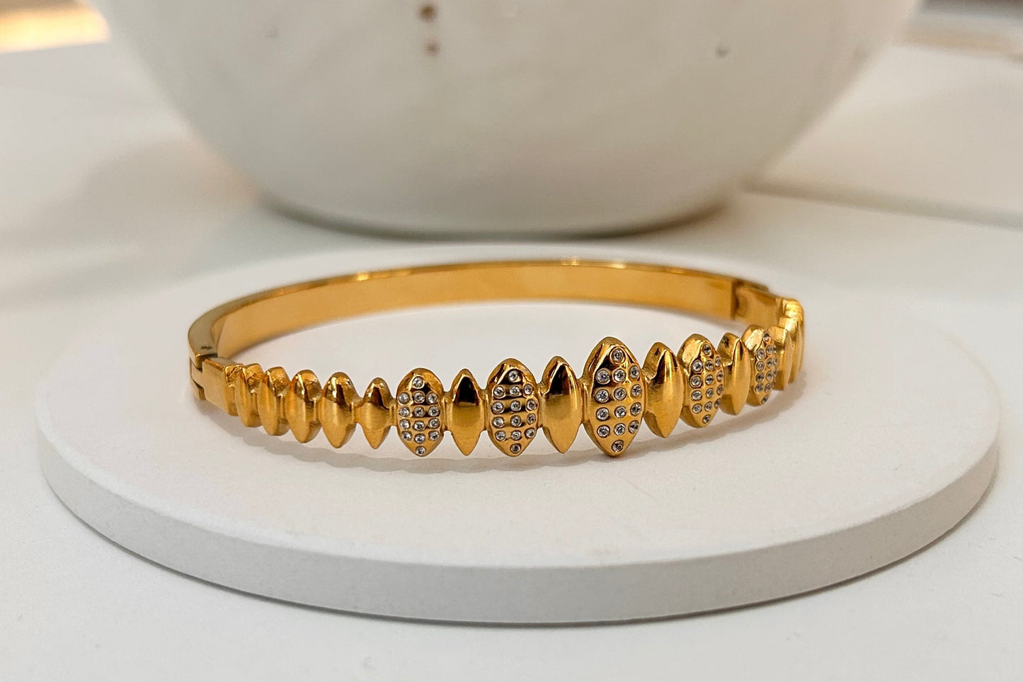 A gold bangle bracelet with a row of decorative pavé and solid teardrop-shaped links, displayed on a round white stand.