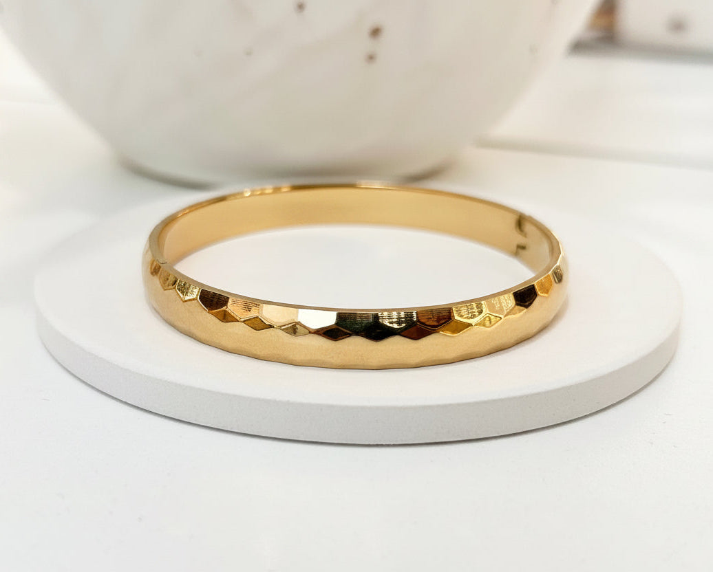 A hammered gold bangle bracelet displayed on a round white coaster, with a blurred white ceramic bowl in the background.