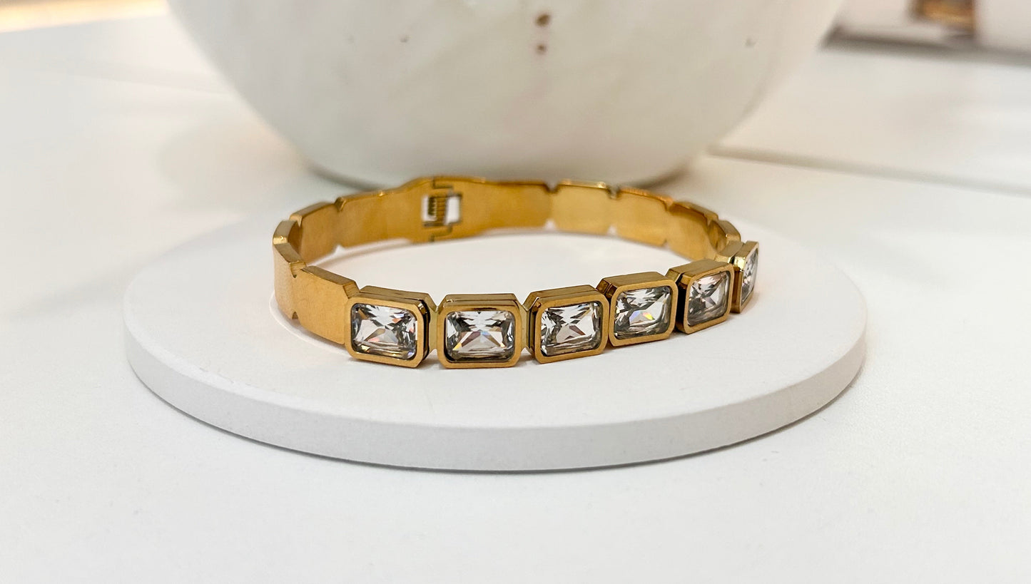 A gold-toned metal bracelet with five rectangular clear rhinestone settings, displayed on a round white pedestal.