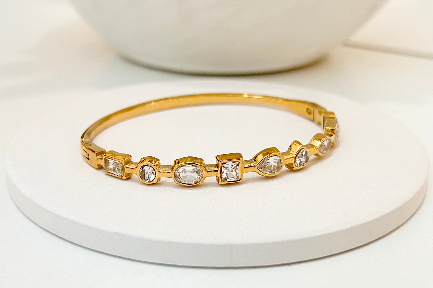 A gold bangle bracelet with five bezel-set clear stones, displayed on a small round white pedestal.