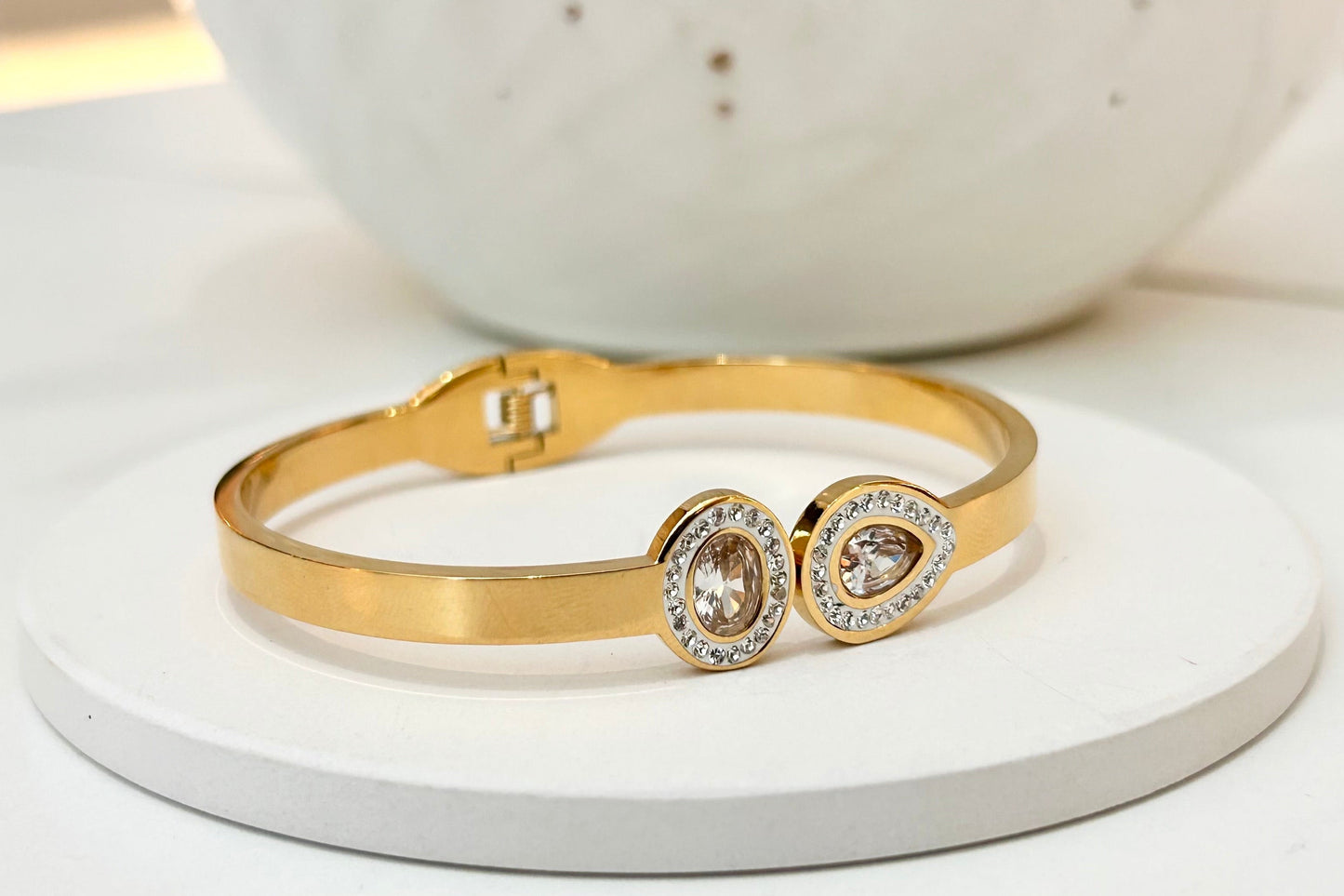 
                  
                    A gold bangle bracelet with two oval bezel-set clear stones, displayed on a round white pedestal with a white speckled ceramic vase in the background.
                  
                