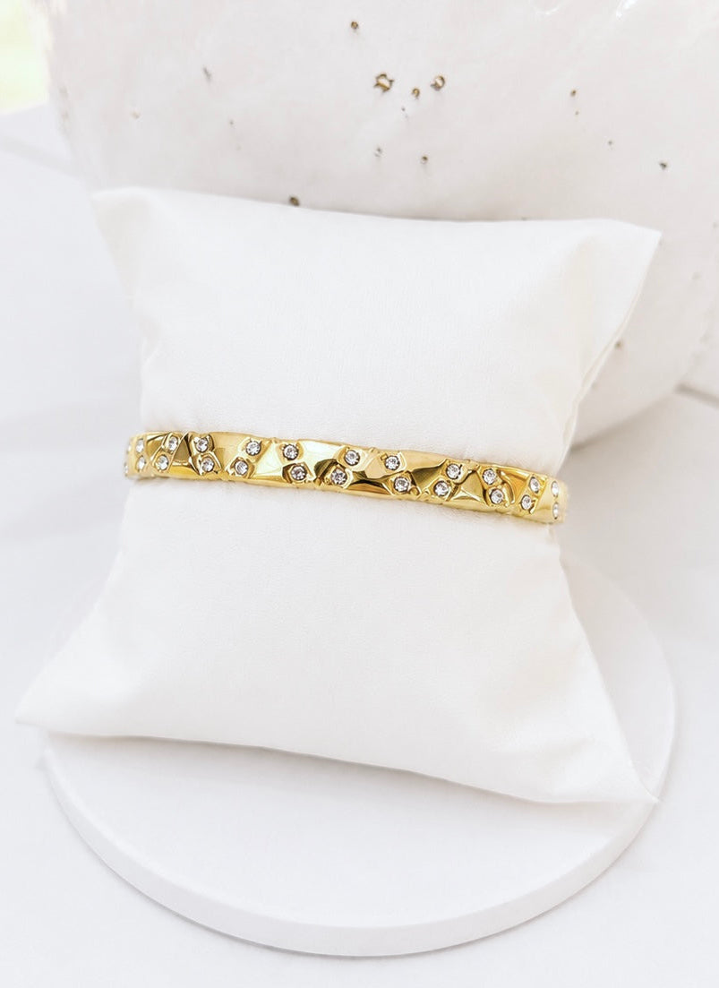 
                  
                    A gold-toned bangle bracelet with small clear rhinestones, displayed on a white fabric jewelry pillow against a bright white background.
                  
                