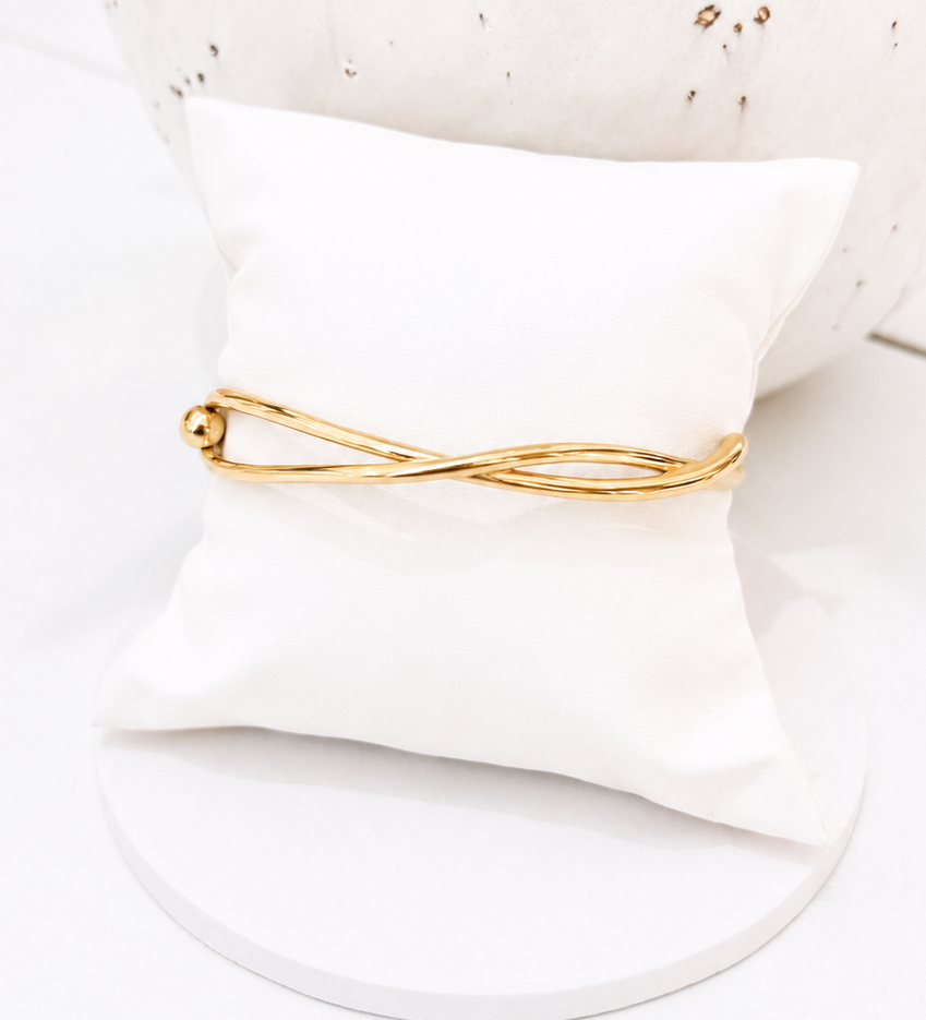 A gold infinity-style ring displayed on a small white pillow, with a large white speckled vase in the background.