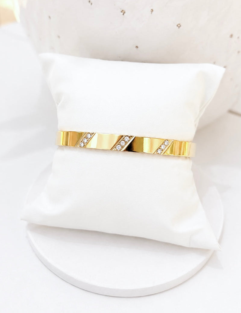 
                  
                    A slim gold bangle bracelet with diagonal crystal-embellished segments, displayed on a small white fabric ring pillow.
                  
                