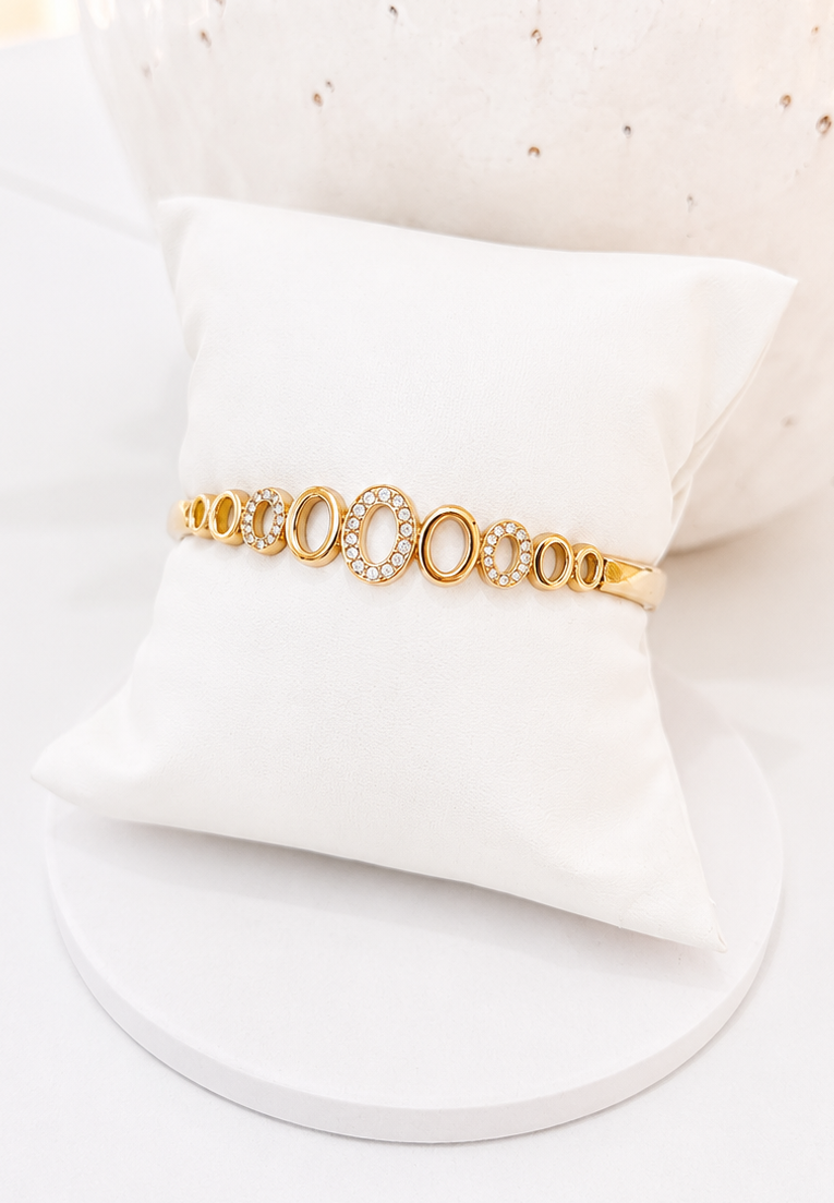 
                  
                    A gold-toned bangle bracelet with oval and circular links, some accented with small clear stones, displayed on a white pillow against a white background.
                  
                