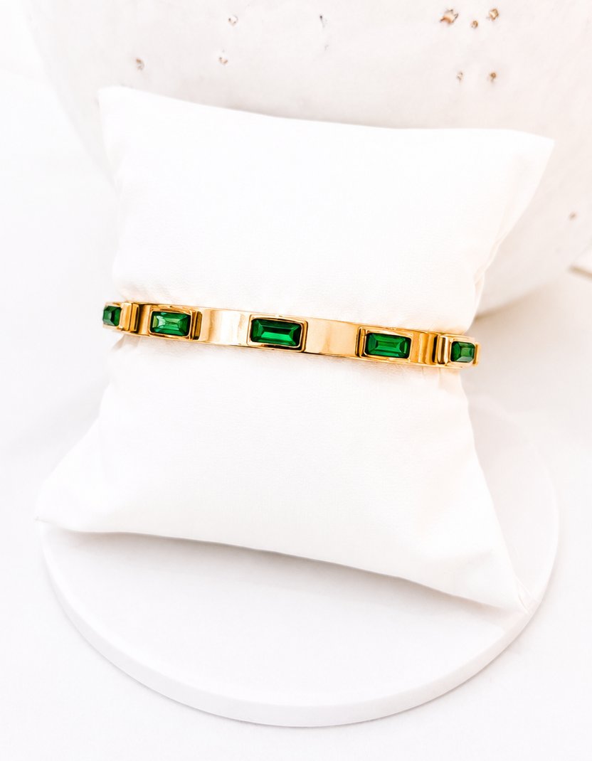 A gold bangle bracelet with rectangular green emerald-cut stones, displayed on a white pillow stand.