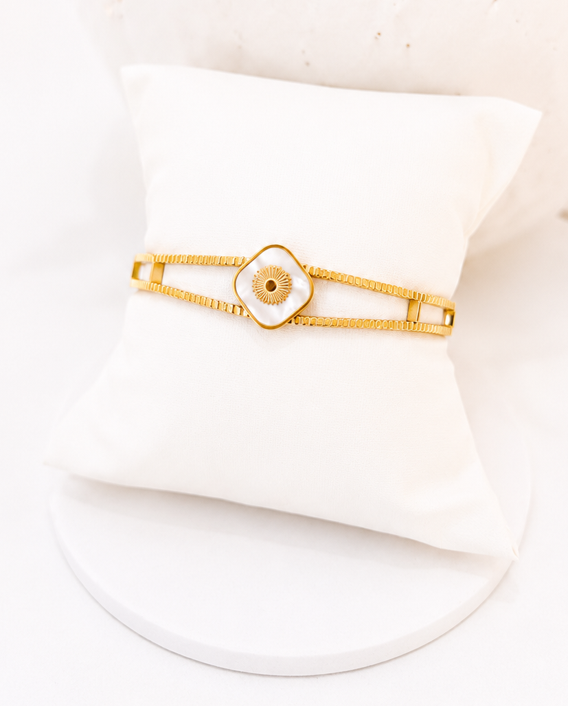 
                  
                    A gold-plated bangle bracelet with a central white enamel lotus flower and a small gold center, displayed on a white cushion.
                  
                