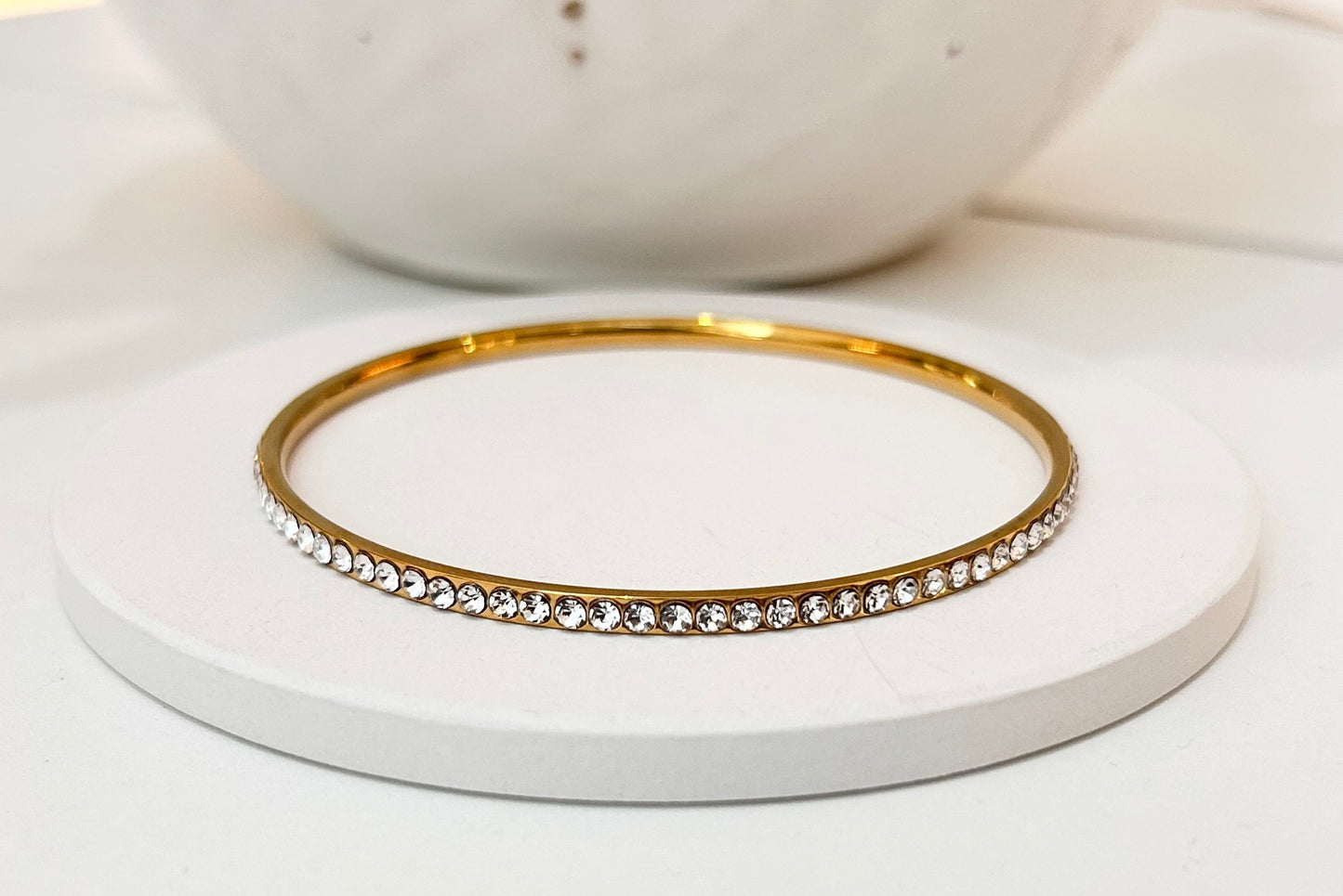 A thin gold bangle bracelet with a row of clear rhinestones, displayed on a round white coaster.