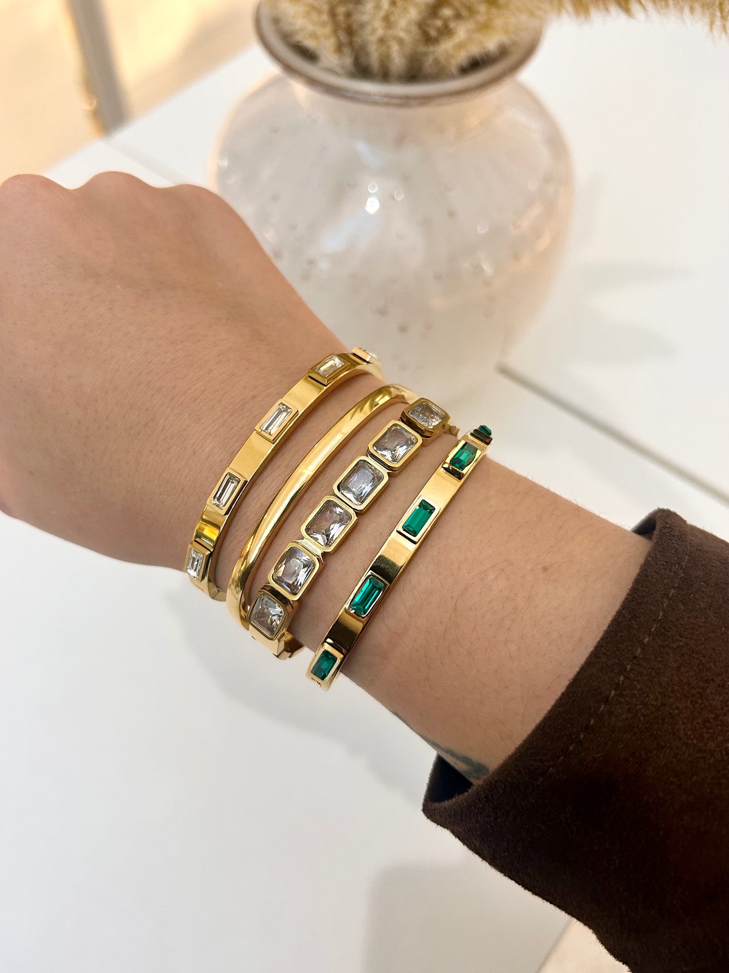 A close-up of a person's wrist wearing four stacked gold bangle bracelets, including some with clear rectangular stones and one with green rectangular stones, against a bright white background with a blurred decorative vase.