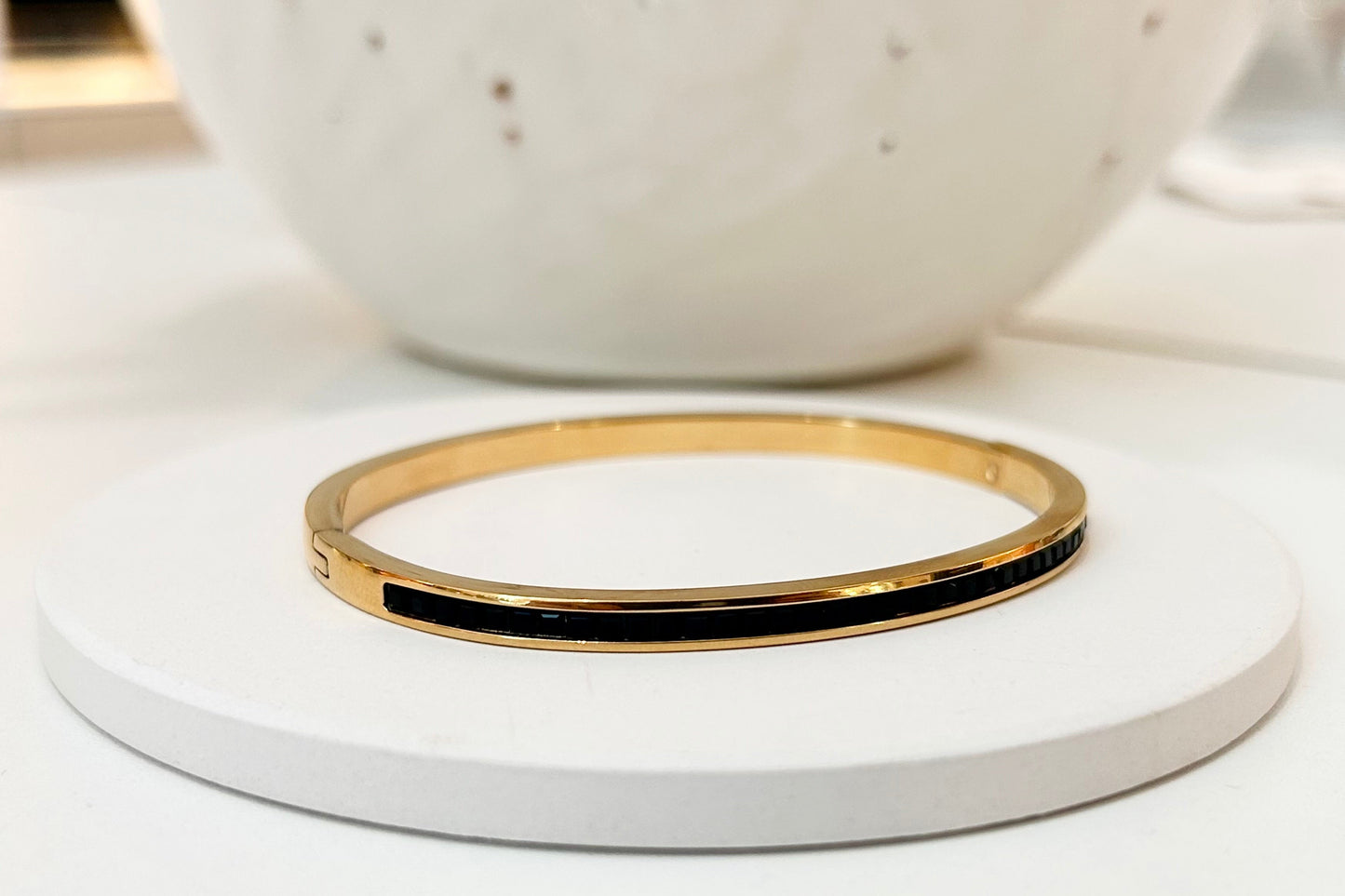 A slim gold bangle bracelet with a black inlay, displayed on a round white coaster with a speckled white ceramic bowl in the blurred background.