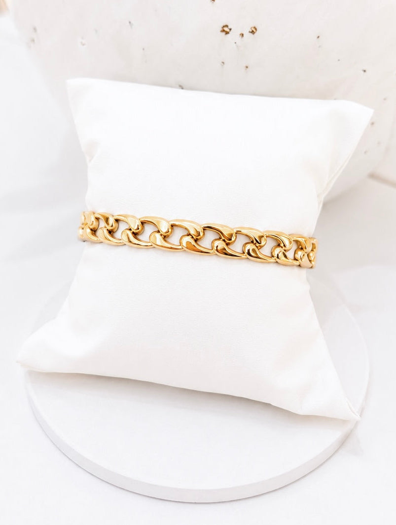
                  
                    A gold chain bracelet displayed on a white fabric jewelry pillow, set against a white background with a decorative ceramic vase.
                  
                