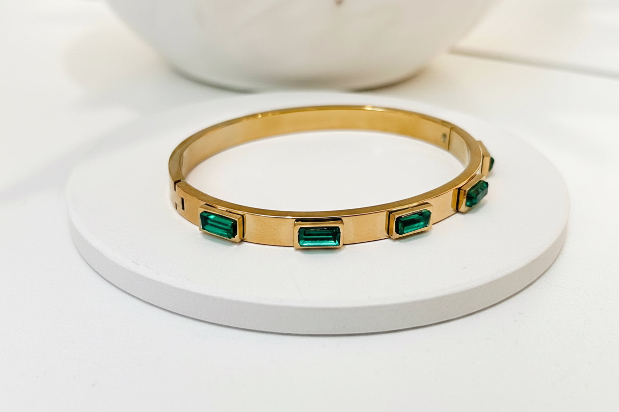 A gold bangle bracelet with rectangular green emerald-cut stones, displayed on a round white pedestal.