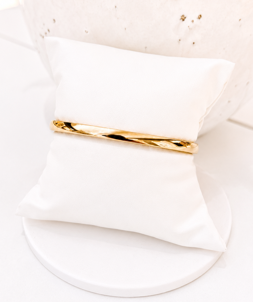 A slim, polished gold band bracelet displayed on a small white fabric ring pillow, set against a clean white background with a textured white vase in the background.