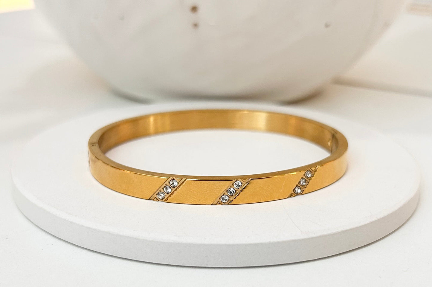 A gold bangle bracelet with two diagonal rows of small clear stones, displayed on a round white coaster with a white ceramic bowl in the background.