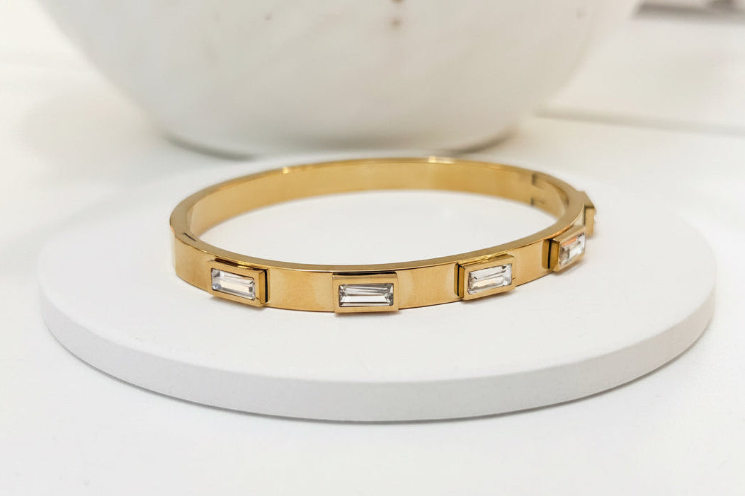 A gold bangle bracelet with rectangular baguette-cut clear stones, displayed on a round white coaster with a white ceramic bowl in the background.