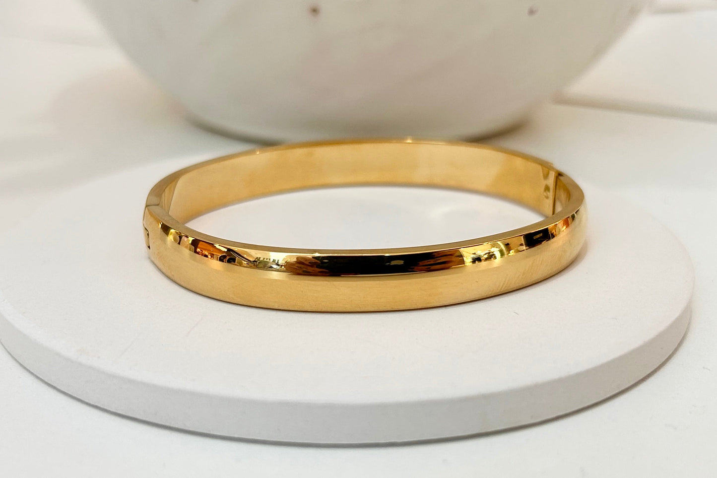 A polished gold bangle bracelet displayed on a round white coaster, with a blurred white ceramic bowl in the background.