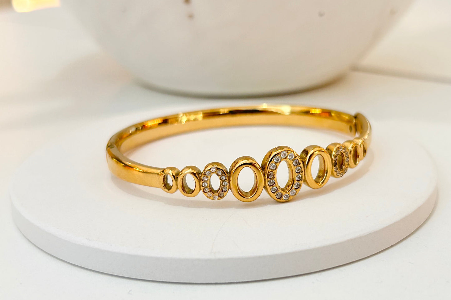 A gold bangle bracelet with a row of oval links, some featuring clear pavé stones, displayed on a white round pedestal.