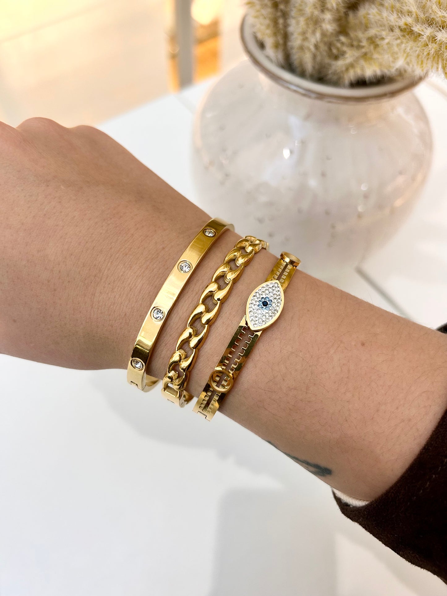 A close-up of a person's wrist wearing four gold-toned bracelets, including a slim tube bracelet with small clear stones, a chunky curb chain bracelet, a textured link bracelet, and an oval evil eye charm bracelet with a blue center.