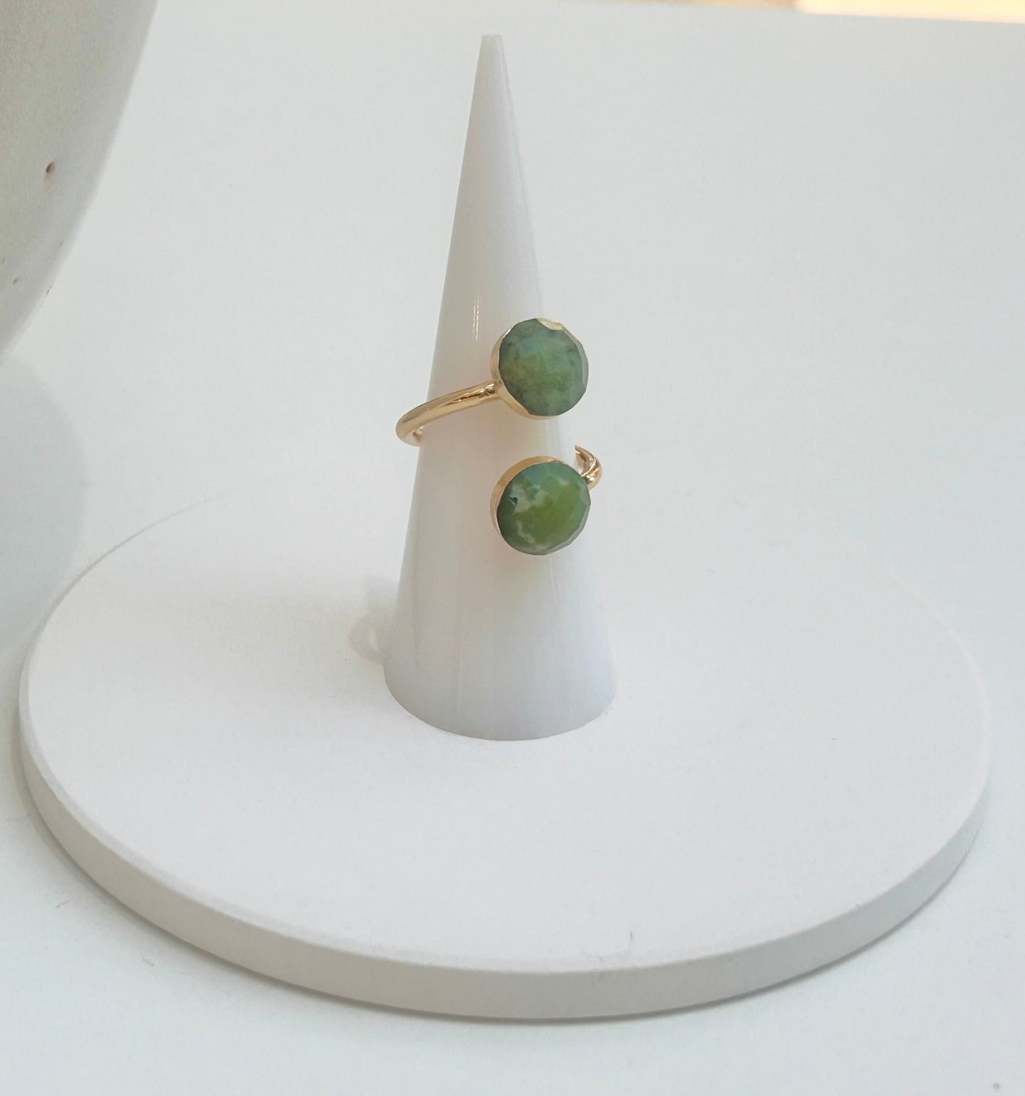 
                  
                    A gold ring with round green jade gemstones displayed on a white cone ring holder, set against a clean white background with a neutral ceramic vessel partially visible on the left.
                  
                
