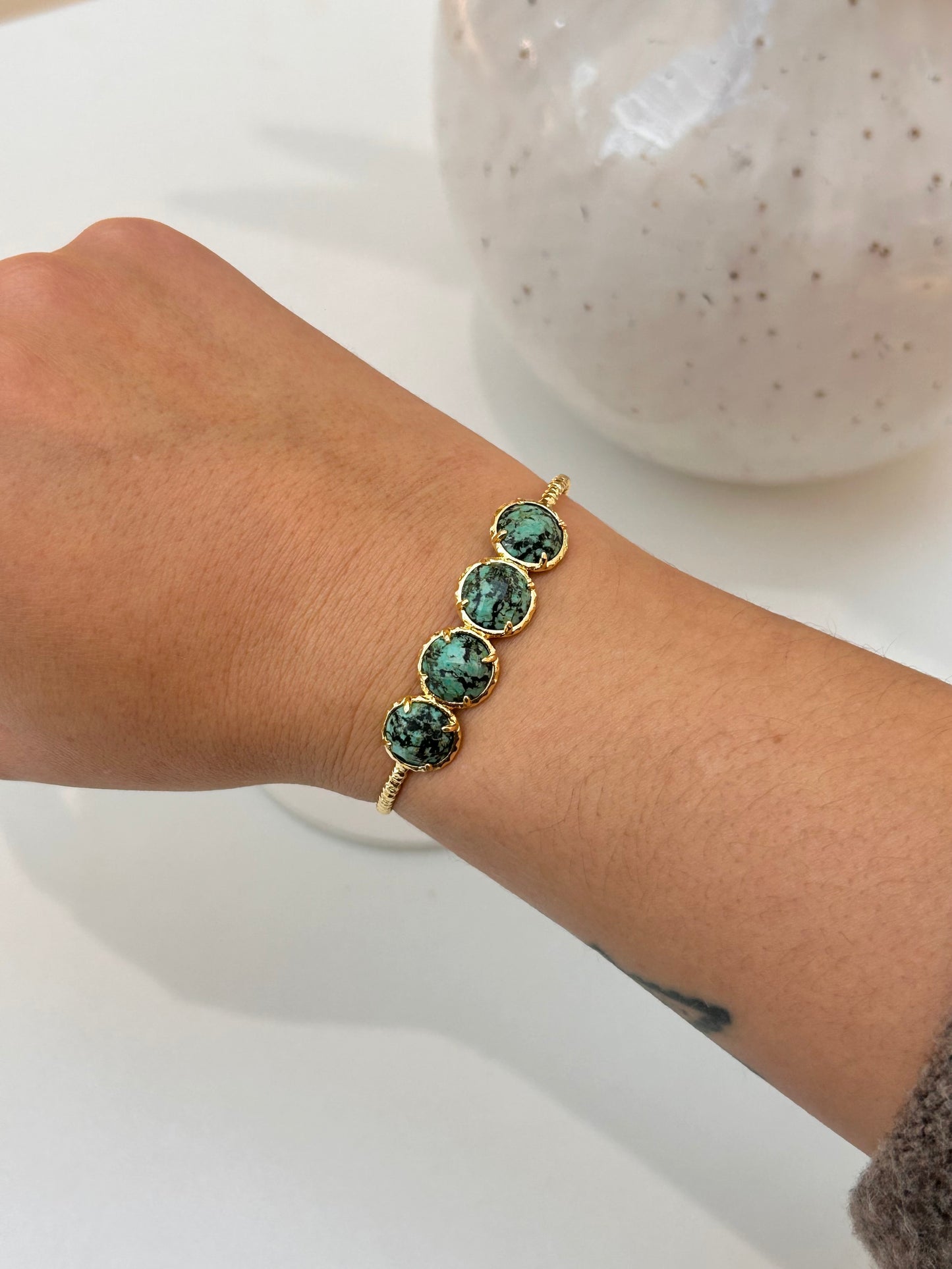 A gold-toned cuff bracelet with four round African Turqouise stones, shown worn on a person's wrist against a white background with a blurred ceramic vase.