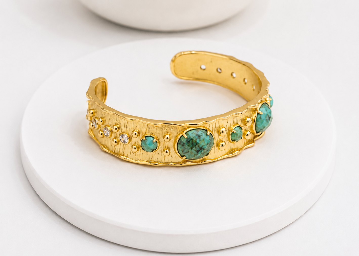 A textured gold cuff bracelet with African turquoise gemstone accents, displayed on a round white pedestal.