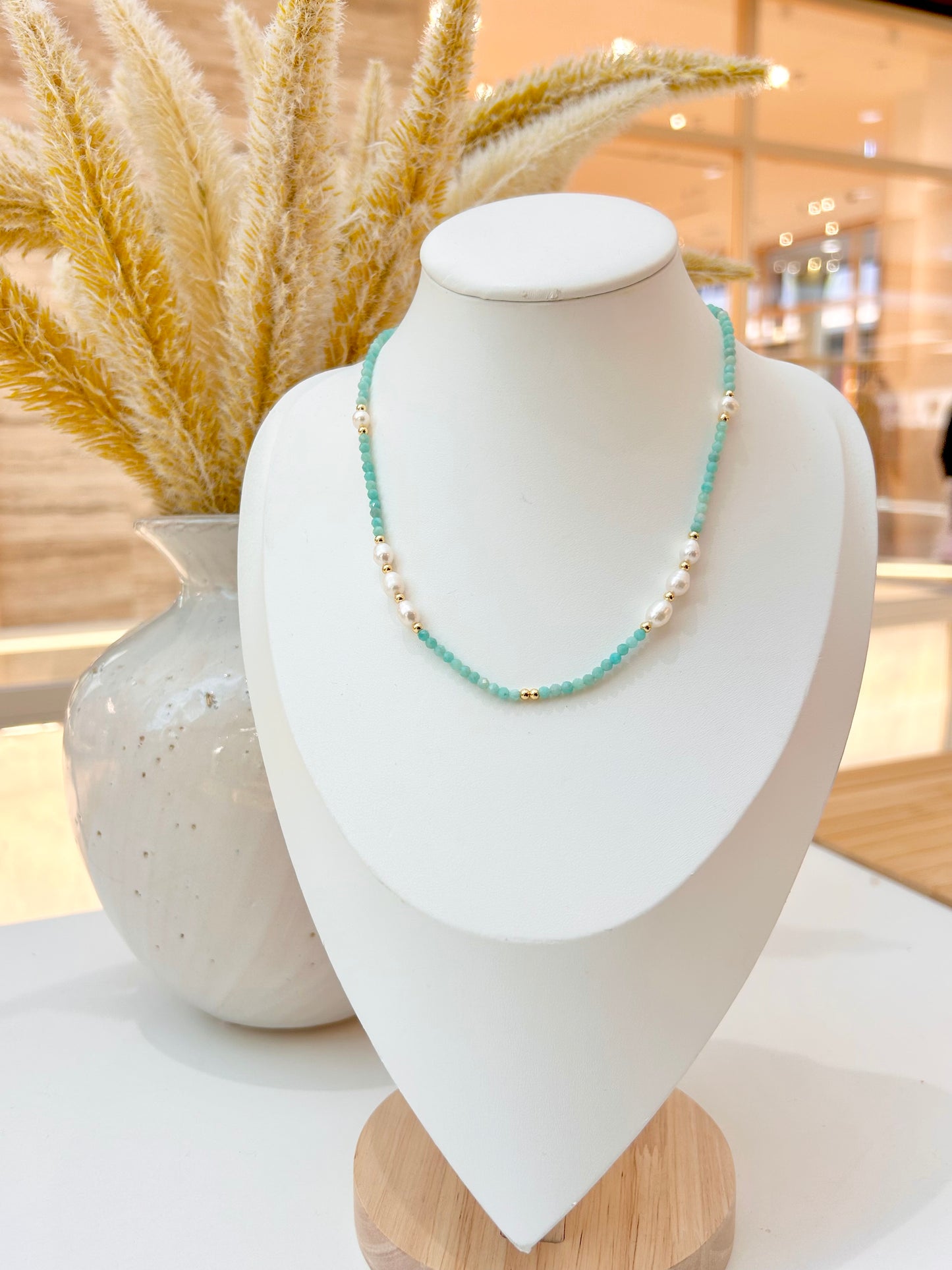 
                  
                    Beaded necklace featuring light amazonite blue-green beads, white pearls, and small gold spacer beads, styled with a cream-colored lace-trimmed top.
                  
                