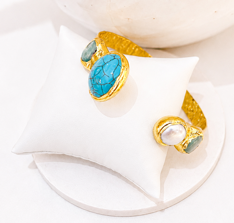 
                  
                    Gold-plated open cuff bracelet featuring natural Amazonite stones and a genuine freshwater pearl, displayed on a white jewellery pillow
                  
                