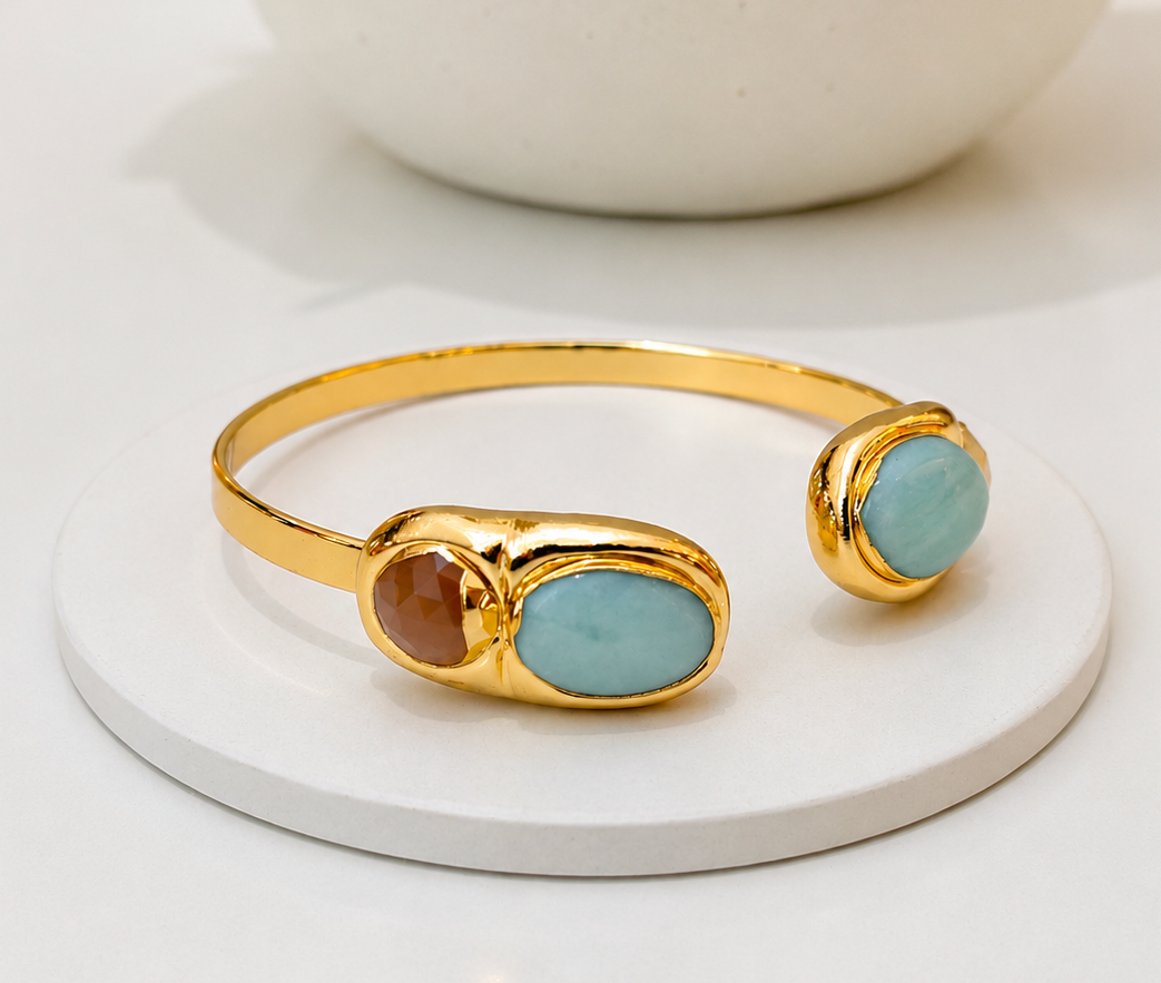 Gold cuff bracelet with two oval light blue amazonite stones and a smaller round sunstone, displayed on a white round coaster.