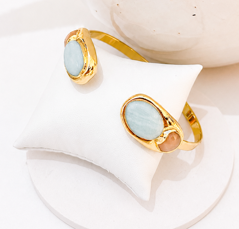 
                  
                    Gold-plated open cuff bracelet featuring smooth natural Amazonite stones and a faceted brown gemstone accent, displayed on a white jewellery stand.
                  
                