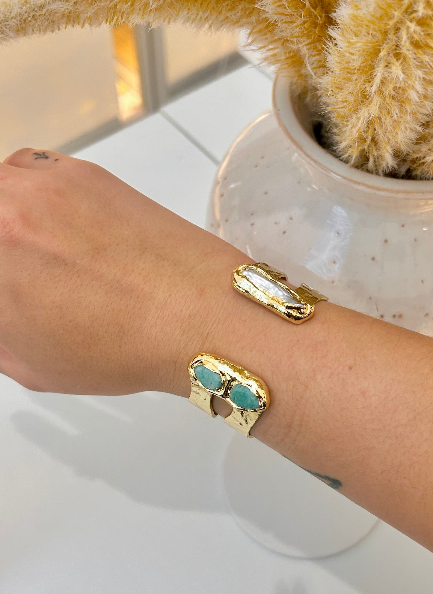 A close-up of a person's forearm wearing a gold cuff bangle with rectangular Amazonite stones and freshwater pearl  inlay details, set against a white tabletop with a ceramic vase in the background.