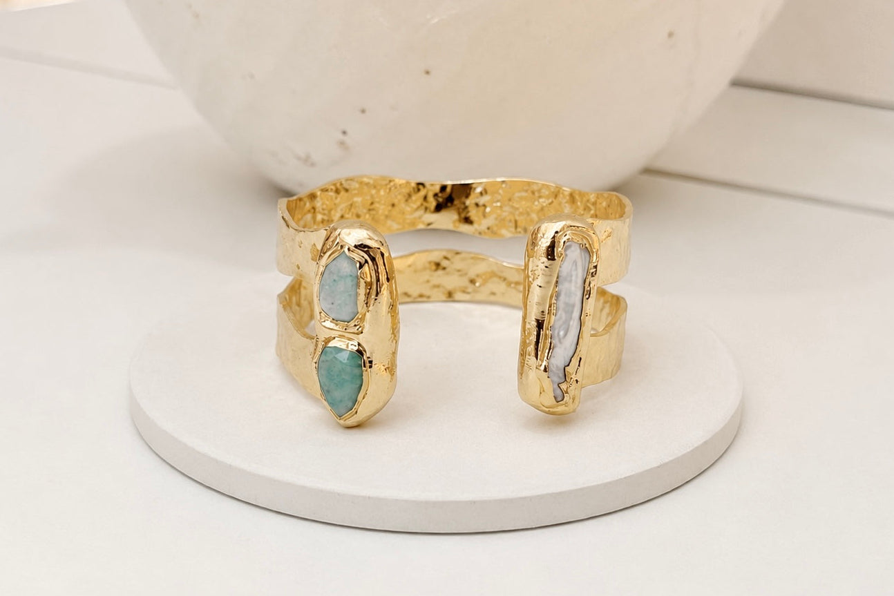 A gold-toned open cuff bracelet with two vertical inlay panels, one amazonite and one freshwater pearl, displayed on a small round white stand.