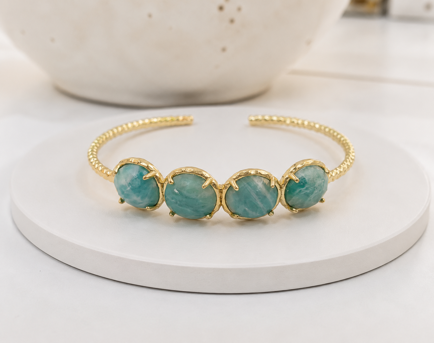 A gold-tone open cuff bracelet featuring four oval amazonite gemstones, displayed on a round white pedestal with a soft-focus ceramic bowl in the background.