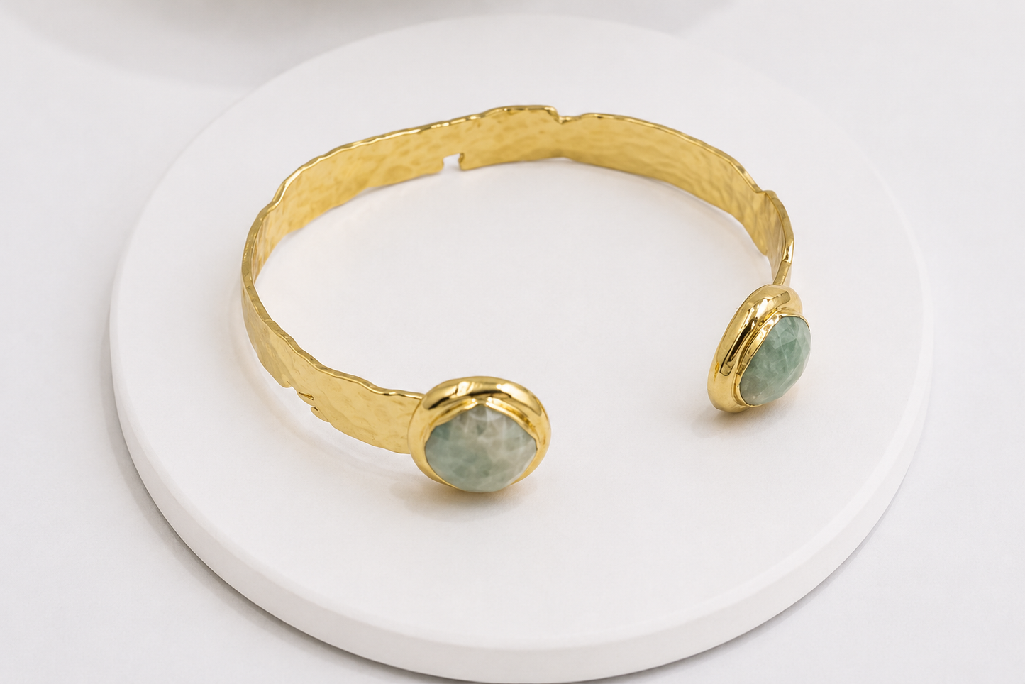 A hammered gold cuff bracelet with two oval amazonite accents, displayed on a round white pedestal.